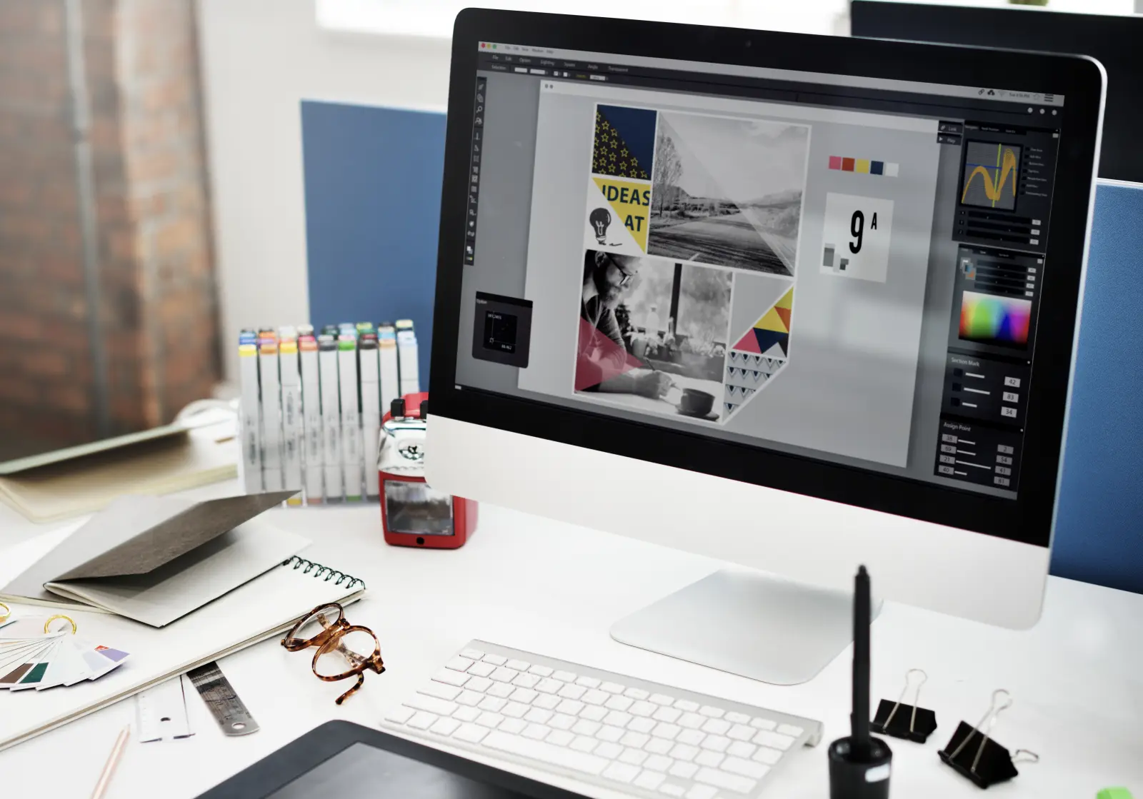 Layar komputer menampilkan software desain berisi moodboard, di meja kerja desainer dengan spidol warna, kacamata, dan contoh palet warna